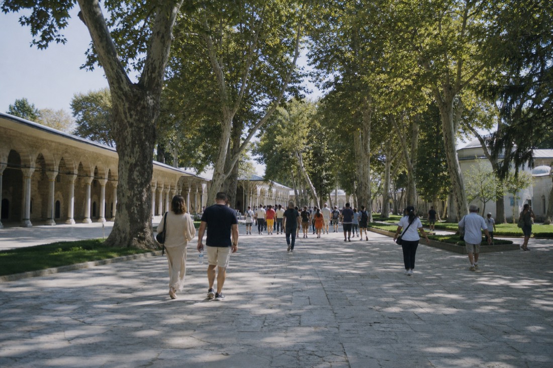 Visitors spread across the tree-lined second courtyard of Topkapı Palace at midday, seen from behind